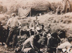 Soldiers, some wounded, some looking over documents, recline or squat in a field