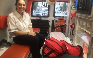 Tess Seltzer rides in the back of an ambulance in a white and black medical uniform, medical equipment in bags at her feet.