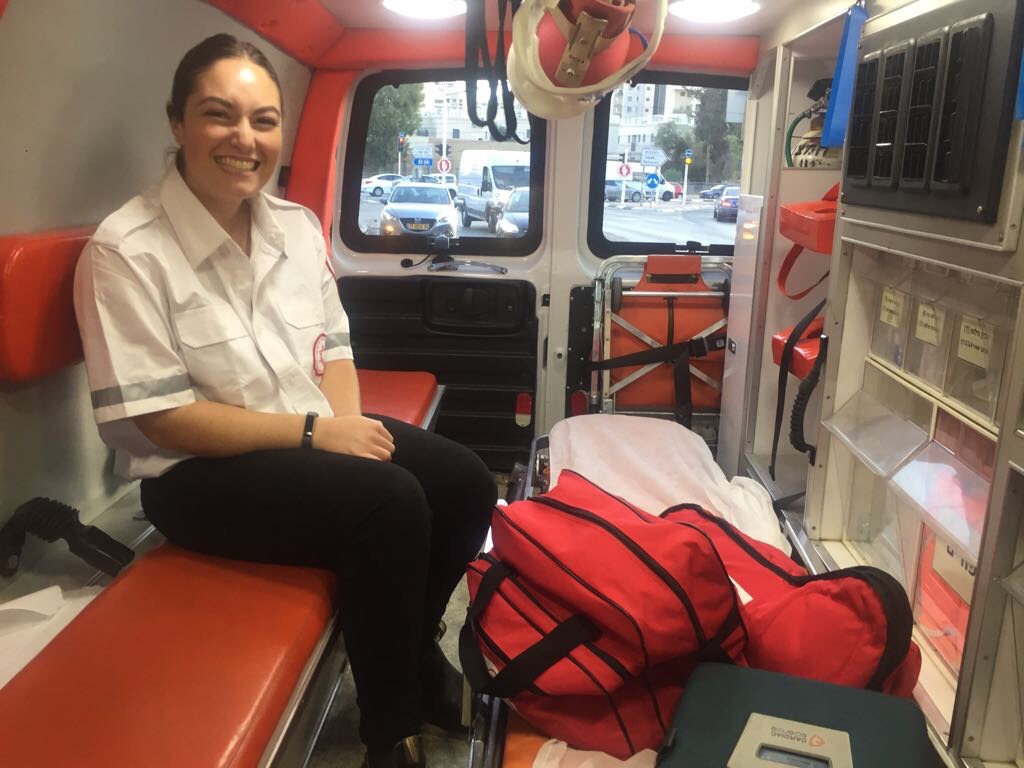 Tess Seltzer rides in the back of an ambulance in a white and black medical uniform, medical equipment in bags at her feet.