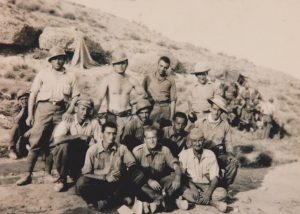 A group of informally dressed soldiers, some wearing helmets, poses for a photo in front of their hillside encampment
