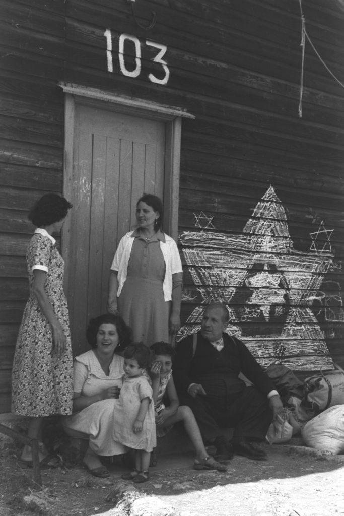 Three women, two children and an elderly man gather, chatting, at the entrance of a large wooden building labeled "103." Stars of David are drawn on the building's wooden exterior in chalk.