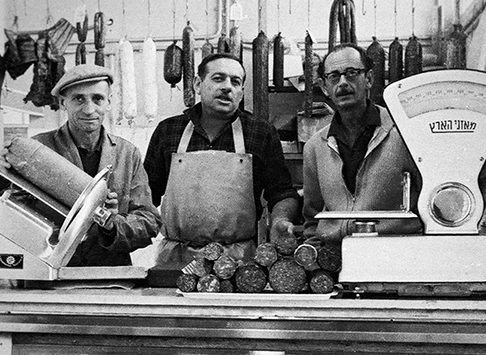 Historical photograph of staff in an Israeli deli, presiding over a pile of salamis