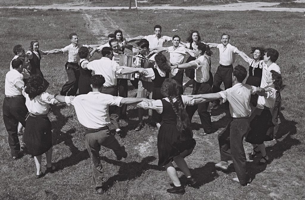 A black and white photograph shows a group of young people dancing the hora in two rings, around accordion-playing musicians on a field 