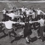 A black and white photograph shows a group of young people dancing the hora in two rings, around accordion-playing musicians on a field