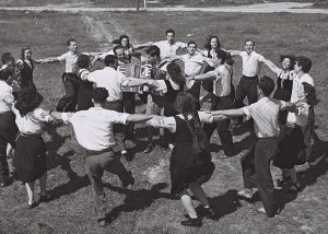 A black and white photograph shows a group of young people dancing the hora in two rings, around accordion-playing musicians on a field