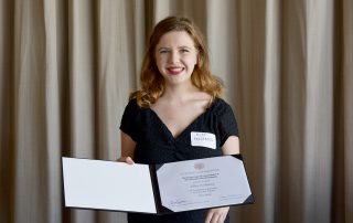 Ellen, wearing a formal black dress, smiles and shows a certificate to the camera