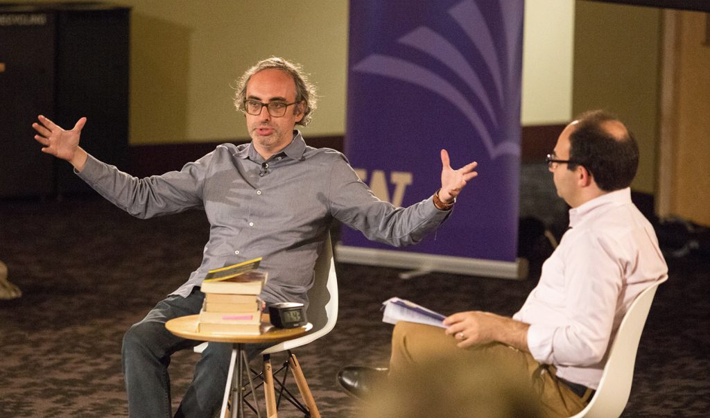 Gary Shteyngart gesticulates, holding his arms wide. He and Professor Sasha Senderovich are seated in the front of Kane Hall at the University of Washington, with a small table stacked with books between them