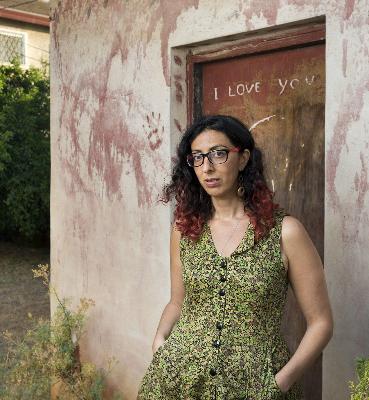 Portrait of Ayelet Tsabari looking serious, hands in pockets of a summer dress, standing in front of the door of a rustic building that has "I love you" scrawled on it