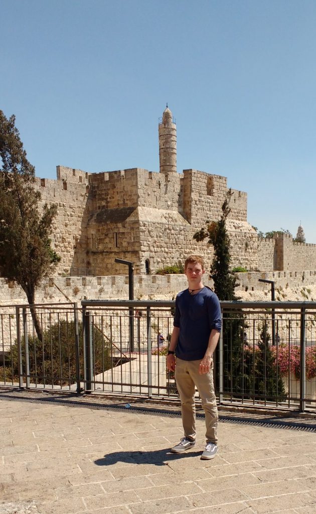 Alex Peterson stands in front of a railing, Jerusalem’s ancient city wall, mosque minaret and blue sky in the background