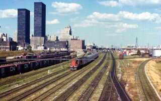 Color photograph shows a train running along rails set up alongside Toronto, identifiable from several skyscrapers and historic large buildings, under blue skies