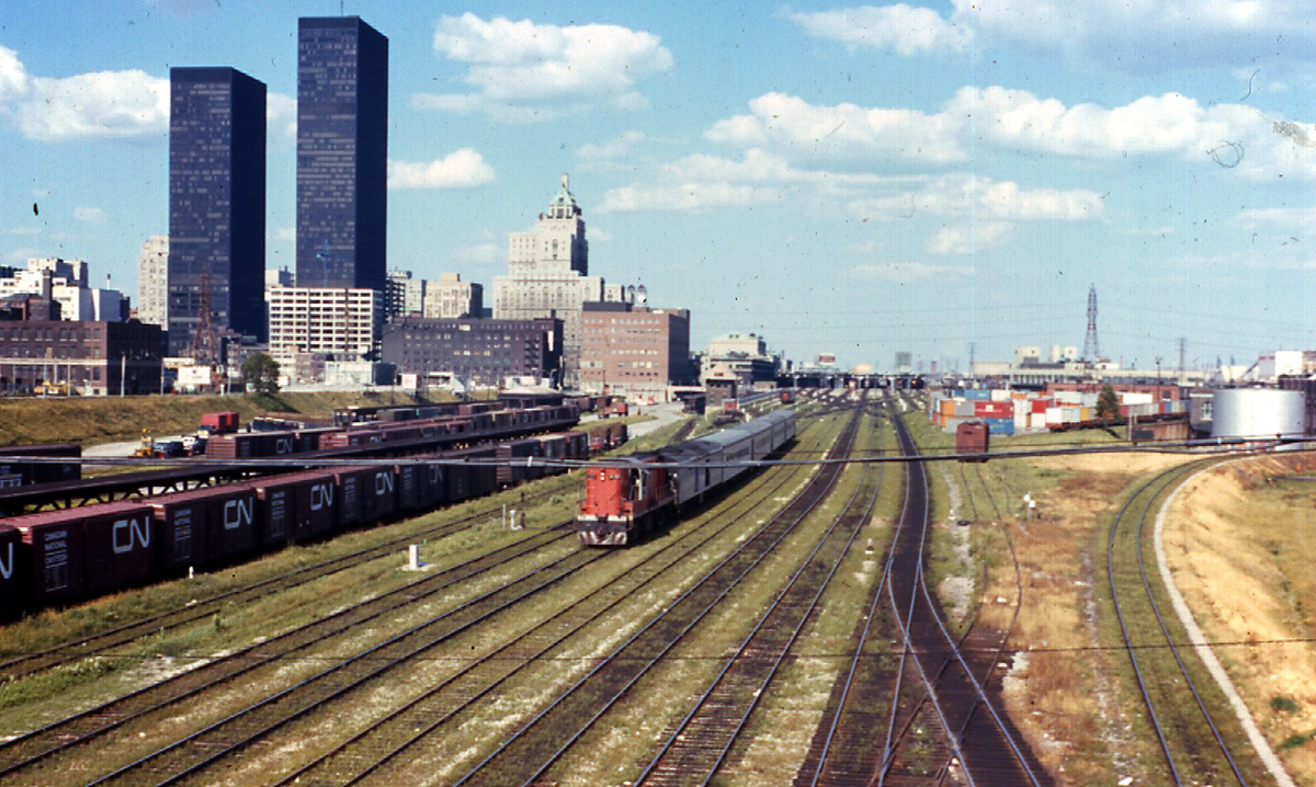 Color photograph shows a train running along rails set up alongside Toronto, identifiable from several skyscrapers and historic large buildings, under blue skies