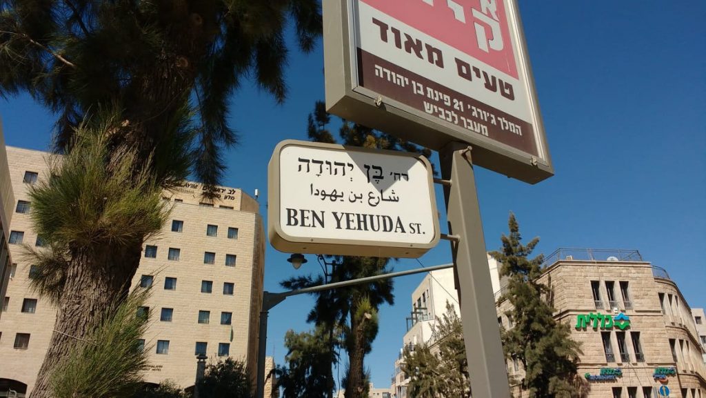 Photograph looking upwards at a blue sky and cream-colored buildings, with palm trees and Hebrew/Arabic store signs on poles in the foreground