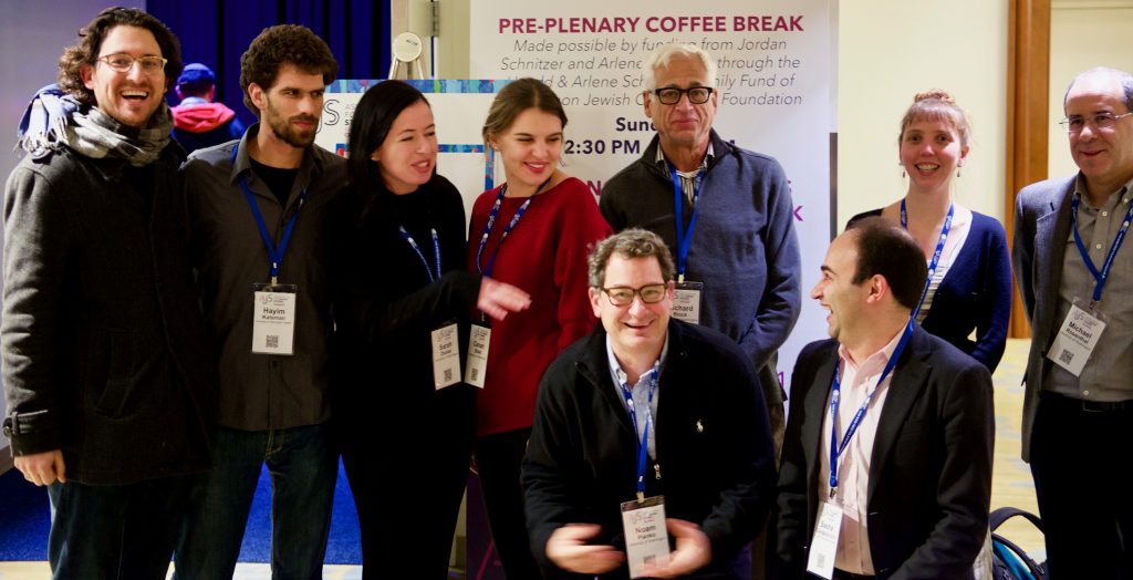 Group photo of several students and faculty members standing or kneeling in a conference center room. Sign in back reads "Pre-plenary coffee break."
