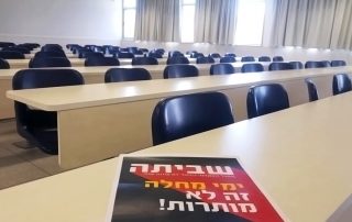 An empty classroom with long tables and seats, all empty. A black and red poster in Hebrew reading "Strike!" sits on the front table.