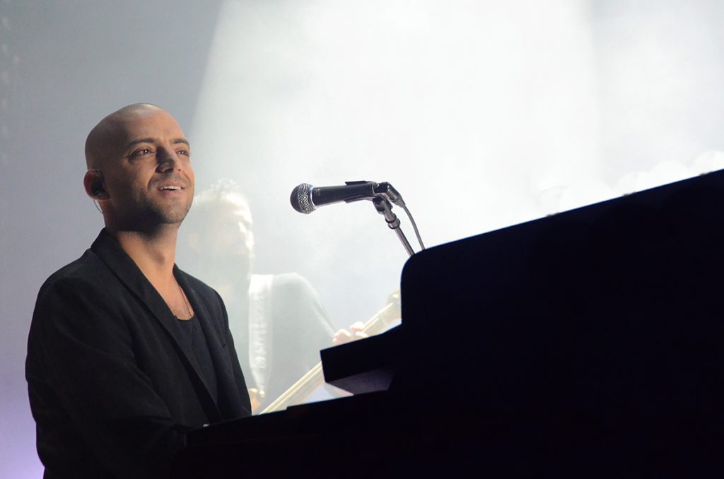 Idan Raichel smiles and looks out at the crowd in concert, playing the piano with a microphone close by.