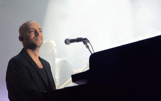 Idan Raichel smiles and looks out at the crowd in concert, playing the piano with a microphone close by.