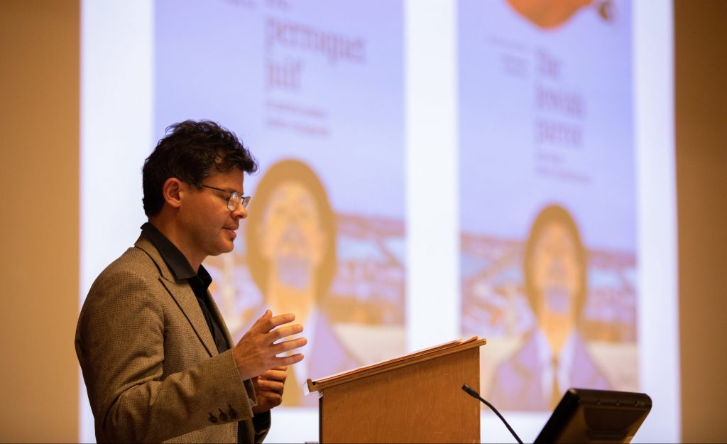 François Azar, wearing a cardigan, button0up shirt, and glasses, speaks at a podium, with the colorful front cover of "The Jewish Parrot" projected in the background