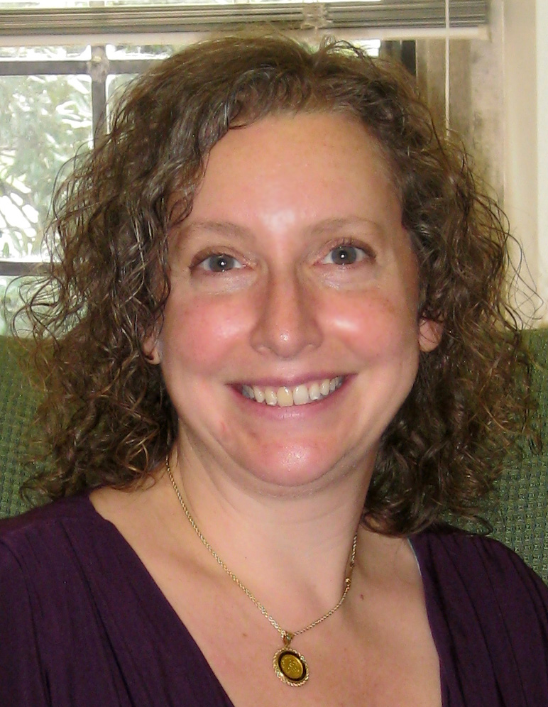 Portrait of Laura Lieber smiling, wearing a purple dress, a window and trees visible in the background