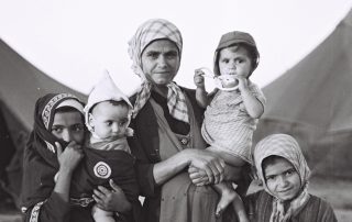 Black-and-white photograph of a woman in a headscarf and long dress holding a baby, with two other girls in scarves and robes, one holding a baby, at her side. Tents are visible in the background.