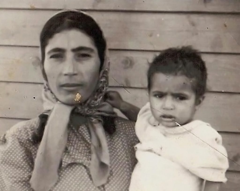Black and white photograph a woman in headscarf and robe holding a baby, slats of a wooden house in the background