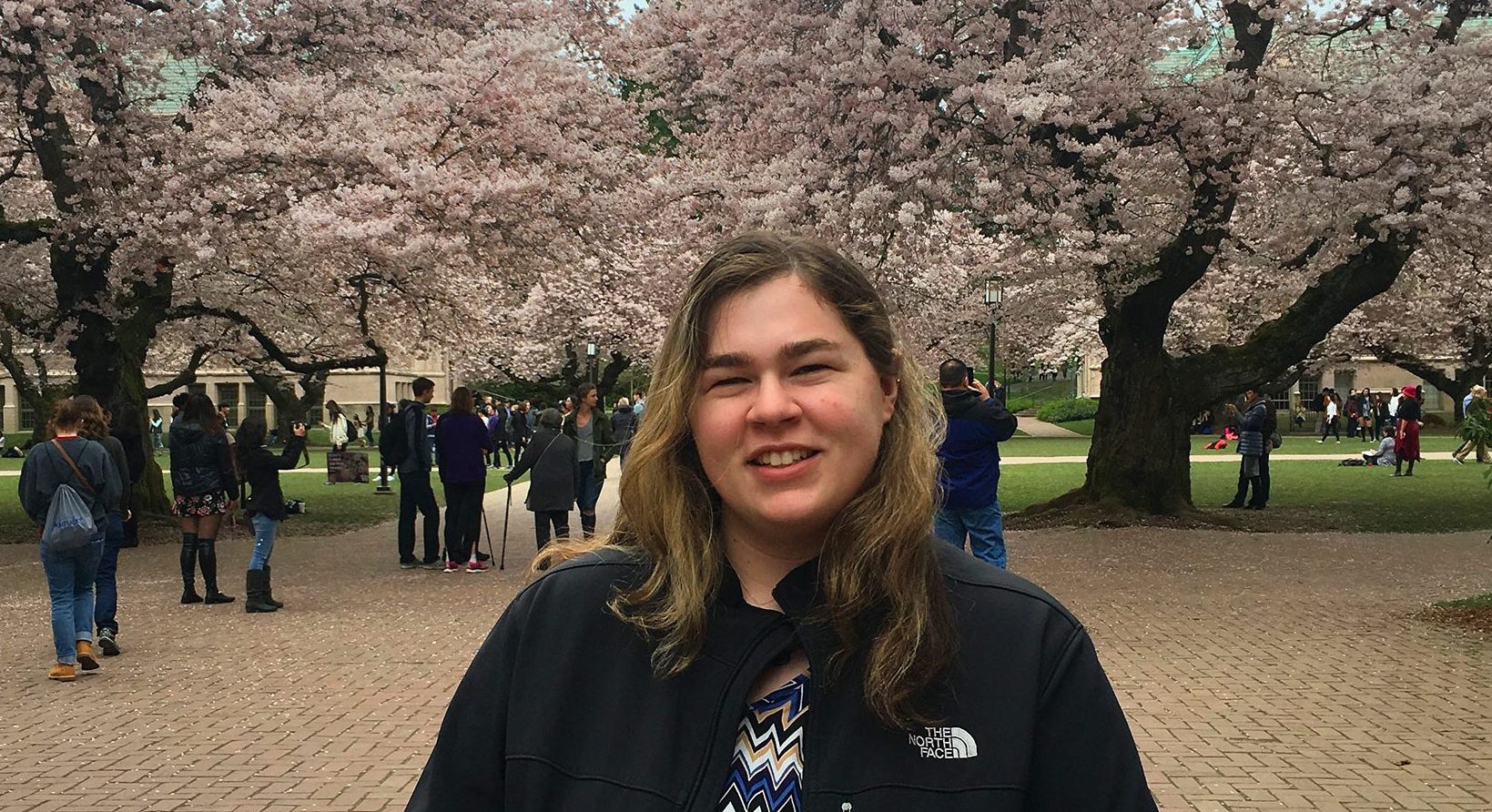 Portrait of Jacqueline Goodrich smiling, in a dark coat and blouse, the UW quad and cherry blossoms in the background