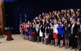 Several dozen employees in formal attire hold their right hands up, taking a pledge onstage in front of Secretary George Pompeo, in suit and tie.