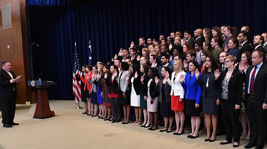Several dozen employees in formal attire hold their right hands up, taking a pledge onstage in front of Secretary George Pompeo, in suit and tie.