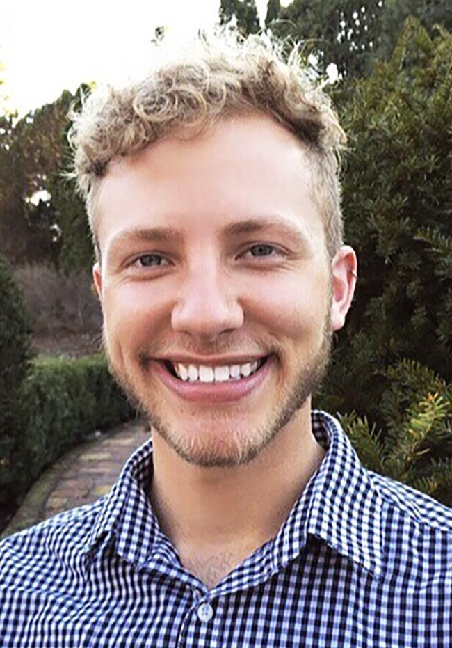 Portrait of Derek Wiebke smiling, wearing a button-up shirt, outdoors with bushes in the background
