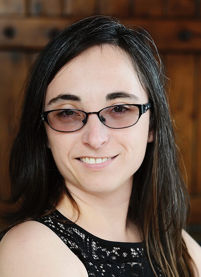 Portrait of Jennifer Hunter smiling, wearing glasses and a dress, with a dark wooden wall in the background