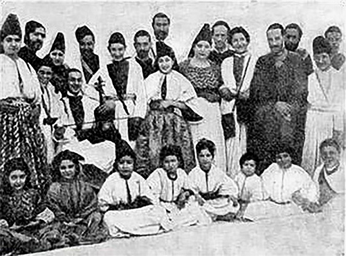 Black and white photograph showing a large group of Moroccan Jews of all ages smiling and wearing traditional clothing: long robes and dresses, shawls, and cylindrical or triangular hats