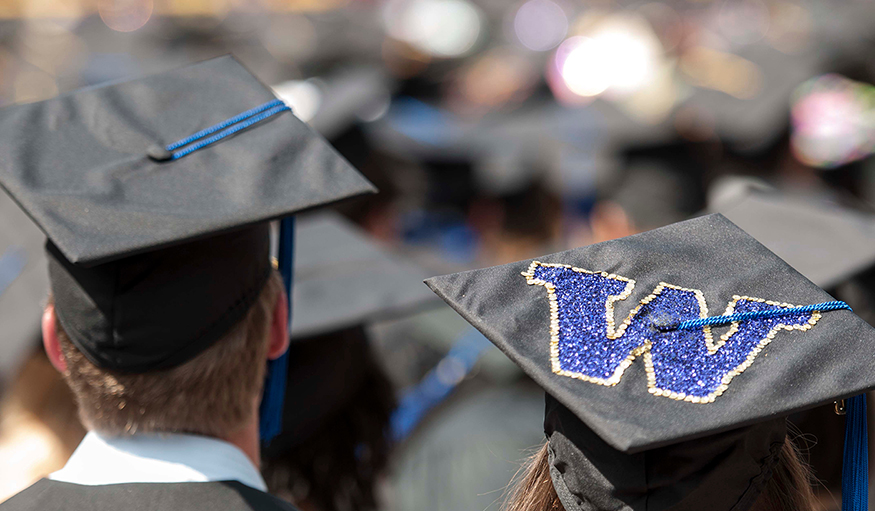 UW graduates stand during commencement, wearing mortarboards that display the purple "W"