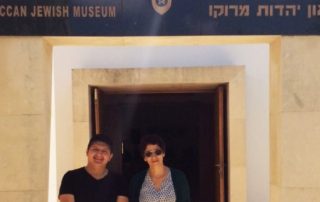 Author Pablo Jairo Tutillo Maldonado in black shirt and pants standing alongside museum curator Zhor Rahihil, wearing blouse, shawl, and khaki pants outside of the museum entrance with sandstone pillars. A black marble marquee reads "Moroccan Jewish Museum" in French, English, Arabic and Hebrew text.