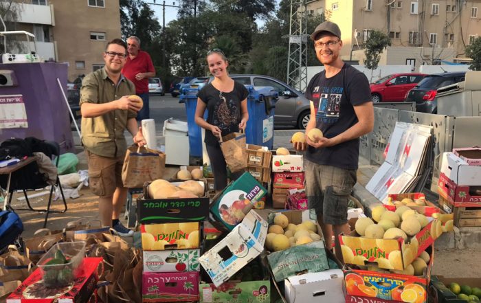 Food frive volunteers, including author Marissa Gaston, stand smiling in front of boxes of produce. A city street, buildings, and trees are visible behind them.