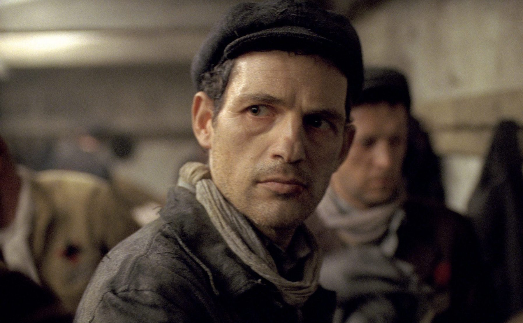 Photograph from the 2015 film "Son of Saul," showing a grim-looking man frowning, wearing a gray hat, scarf, and heavy coat