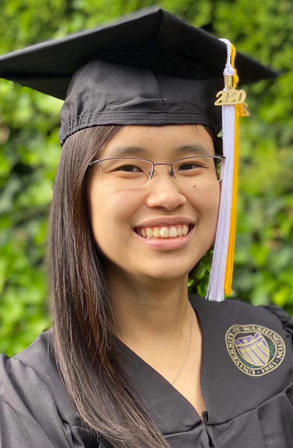 Portrait of Grace Dy, smiling, wearing a black graduation robe a cap with white and gold tassels, with green ivy visible in the background