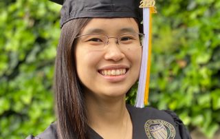 Portrait of Grace Dy smiling, in glasses, wearing a black square graduation cap (mortarboard) and gown, with a white and gold tassel. A verdant hedge is visible in the background.