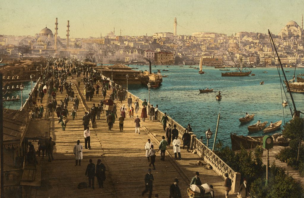 A colorized photograph shows a wide wooden bridge spanning a boat-filled river in Istanbul. Dozens of men wearing fez hats and long coats walk along the bridge. A dense cityscape and several large mosques are visible in the background.