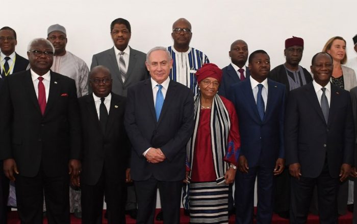 Wide-angle photograph showing Benjamin Natanyahu standing among West African leaders, all wearing formal attire, with a white wall and banners in the background