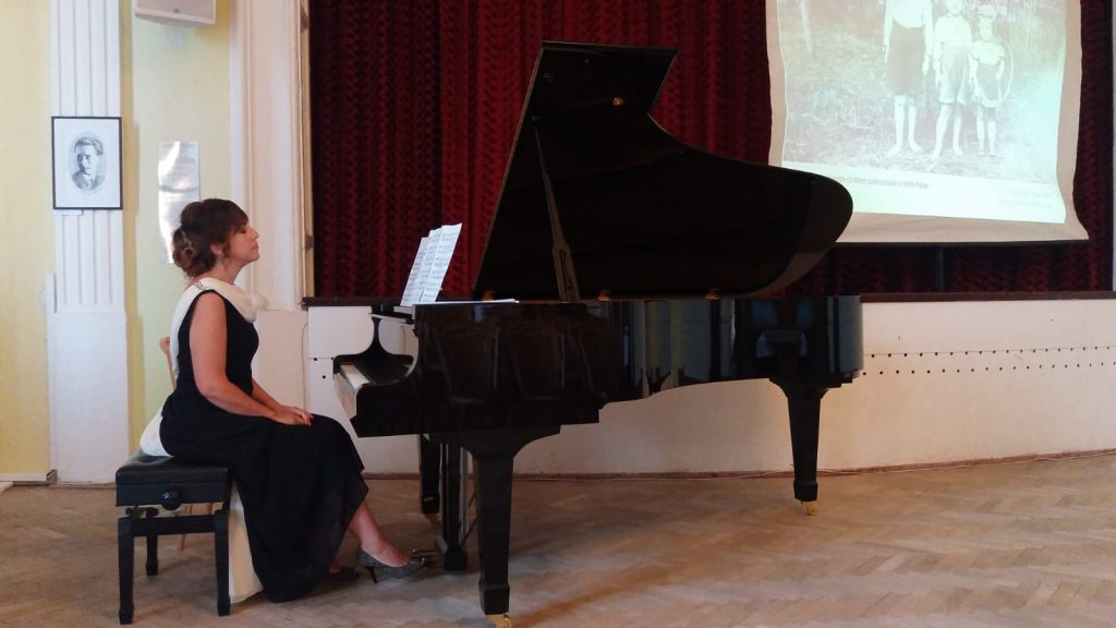 Renan Koen seated at a black grand piano. Renan is wearing a black sleeveless dress and is sitting straight-backed at the piano with hands on her lap. The room has a light brown wood floor and a burgundy wall with white trim around the baseboard and windows. 
