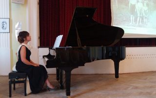 Renan Koen seated at a black grand piano. Renan is wearing a black sleeveless dress and is sitting straight-backed at the piano with hands on her lap. The room has a light brown wood floor and a burgundy wall with white trim around the baseboard and windows.