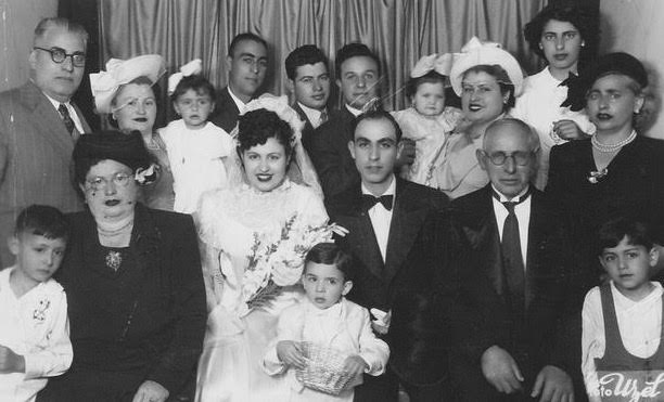 Black and white family photo of a wedding in Tekirdag, Turkey. Bride and groom seated middle with many family members around them in formal attire. Child seated on bride's lap. Background is a panel of curtains.