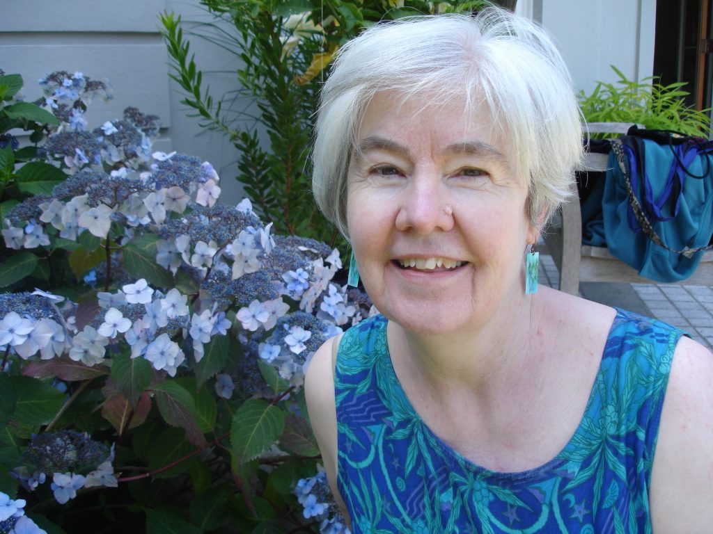 Headshot of Maureen Jackson. She has short white hair and is wearing a blue and purple tank top and blue earrings. In the background there is a purple and blue hydrangea bush. She is smiling.