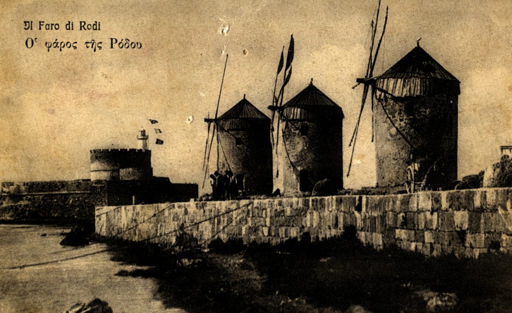 Old sepia toned postcard from Rhodes. Features three brick windmills in the foreground and a brick wall along a sea line.