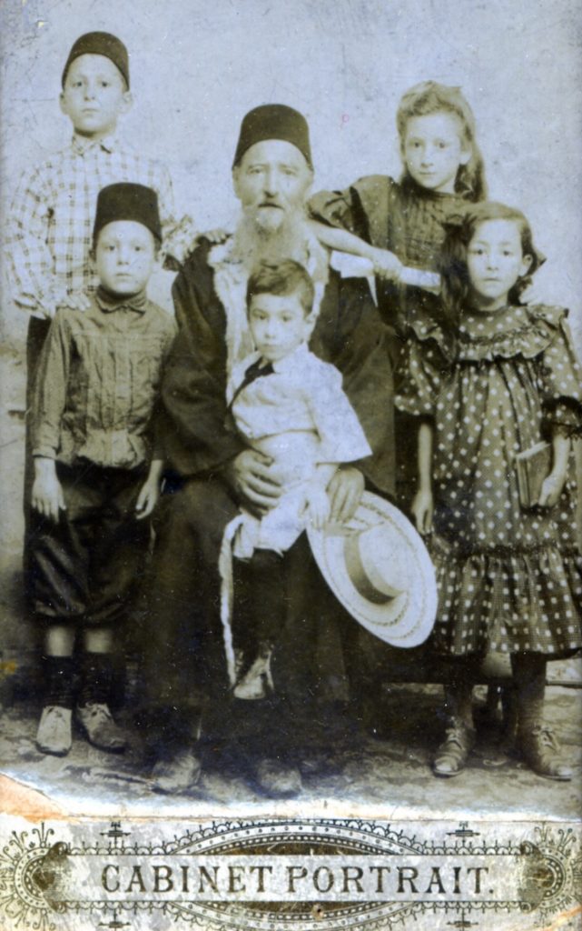 Old family portrait in sepia tone. Father seated in middle wearing a fez hat and robe with a child on his lap, who is holding a ladies bolero hat. Around the father are his children, two boys at left and 2 girls at right, all in 1920s attire. The bottom of the photo has a printer's stamp that says Cabinet Portrait.