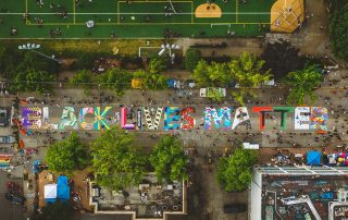 Overhead photograph showing a colorful "Black Lives Matter" mural painted on a street next to a grassy park. Photo by Kyle Kotajarvi.