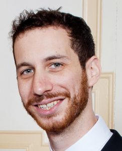 Portrait of Brendan Goldman, smiling, wearing a collared button-up white shirt and black cardigan, standing with a collegiate-looking wall in the background