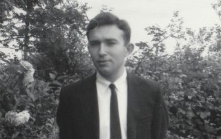 Black and white photograph shows a young Edward Alexander, wearing a button-up shirt, tie and suit jacket, standing in front of greenery, looking contemplative