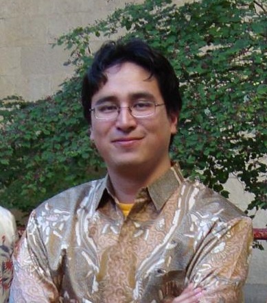 Headshot of Jorge. He is wearing a tan colored button down shirt. Jorge has glasses and black wavy hair. His arms are crossed and he is smiling with his mouth closed. There is a wall behind him with green ivy.