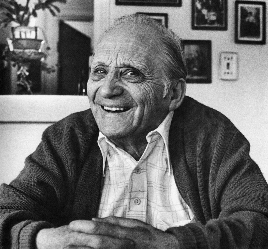 Black and white portrait of Henry Benezra wearing a button down and sweater. Framed photos in background.