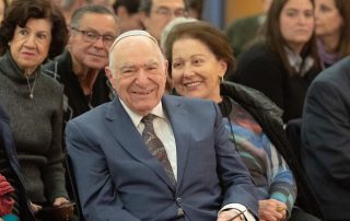 Hazzan Azose smiling in the crowd at Ladnio Day wearing a grey suit, white shirt, and tie. His wife Elisa is next to him.